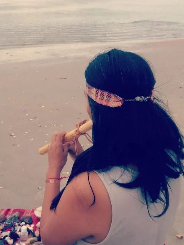 Mujer tocando una flauta en la playa, con el mar al fondo, sentada sobre una manta decorativa.