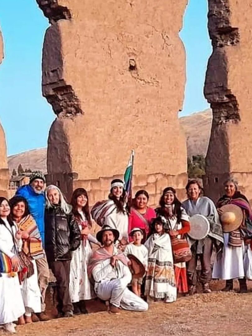 Grupo de personas vestidas con ropa tradicional frente a estructuras de piedra en un paisaje andino, participando en una ceremonia o ritual.
